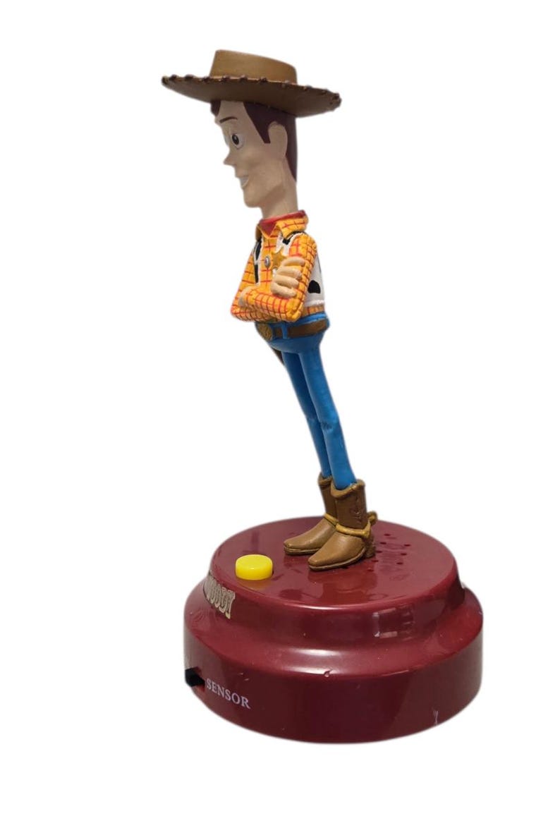 May include: A Woody action figure from the Toy Story franchise. The figure is wearing a cowboy hat, plaid shirt, blue jeans, and brown boots. The figure is attached to a red base with a yellow button.