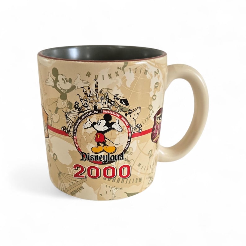 Pode incluir: Caneca de cer&acirc;mica bege com Mickey Mouse e o logotipo da Disneyland 2000. A caneca tem uma al&ccedil;a creme e um interior cinza escuro. O design inclui um mapa-m&uacute;ndi e v&aacute;rios &iacute;cones da Disneyland.