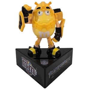 May include: A yellow M&M's candy character in a Bumblebee Transformer costume stands on a black triangular base. The base features the M&M's logo and the Transformers logo. The figure has black and yellow accents.