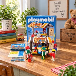 Puede incluir: Un set de juguetes Playmobil que incluye un puesto de mercado con toldo a rayas, varios artículos de comida en miniatura y cuatro figuras pequeñas. El set contiene una bicicleta roja, una caja azul con imágenes y un manual sobre madera.
