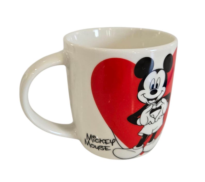 Op de afbeelding: Witte keramische mok met een gebogen handvat, met Mickey Mouse voor een groot rood hart. De woorden "Mickey Mouse" staan op de zijkant gedrukt. Een klassiek Disney-ontwerp.