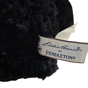 May include: Close-up of a black, textured fabric with a tag that reads "Eddie Bauer by Pendleton". The tag is rectangular with a cream background and dark blue text. The fabric appears soft and plush.