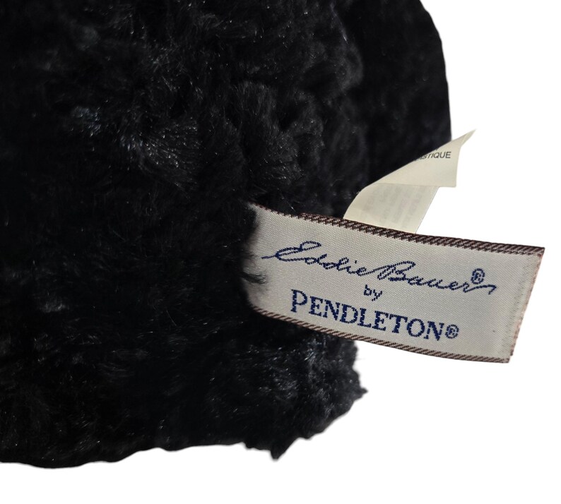 May include: Close-up of a black, textured fabric with a tag that reads "Eddie Bauer by Pendleton". The tag is rectangular with a cream background and dark blue text. The fabric appears soft and plush.