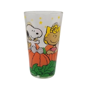 May include: A clear glass featuring Snoopy and Sally Brown from Peanuts, sitting on pumpkins. The glass has a white background with yellow stars. Green leaves are at the bottom. A fun, decorative glass for fans of the comic strip.