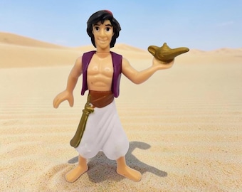 May include: A plastic figurine of a character with black hair, wearing a purple vest, a brown belt, and white pants, holding a small gold-colored lamp in one hand. The figure has a tan skin tone and is standing upright with bare feet.