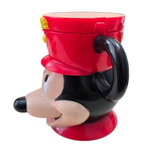 May include: A novelty mug shaped like Mickey Mouse in a red and black marching band uniform. The mug features a black handle and a red hat with yellow accents. The mug is made of a glossy material and has a detailed design.