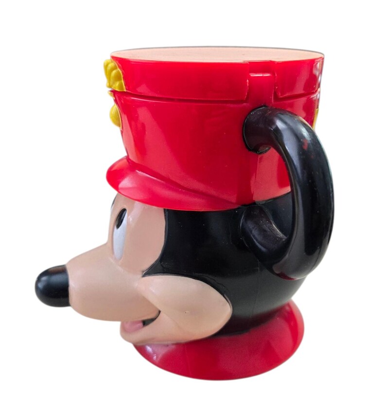 May include: A novelty mug shaped like Mickey Mouse in a red and black marching band uniform. The mug features a black handle and a red hat with yellow accents. The mug is made of a glossy material and has a detailed design.