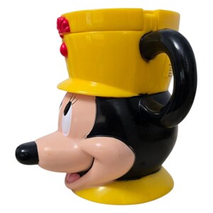 May include: A yellow and black Minnie Mouse-shaped mug with a black handle. The mug features Minnie's face, with a yellow hat and red accents. The mug is made of a glossy material and is designed for beverages.