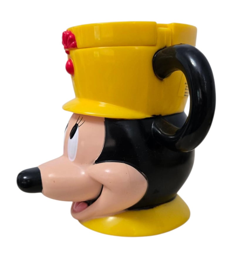 May include: A yellow and black Minnie Mouse-shaped mug with a black handle. The mug features Minnie's face, with a yellow hat and red accents. The mug is made of a glossy material and is designed for beverages.