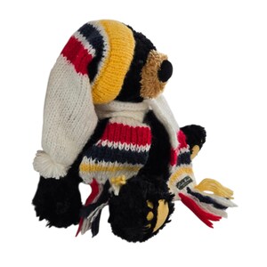 May include: A black plush bear wearing a knit hat and scarf. The hat is white, yellow, red, and navy blue. The scarf has matching colors and a small tag. The bear has a tan muzzle and yellow paw pads.