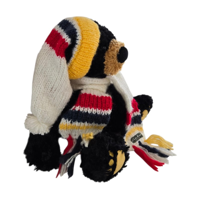 May include: A black plush bear wearing a knit hat and scarf. The hat is white, yellow, red, and navy blue. The scarf has matching colors and a small tag. The bear has a tan muzzle and yellow paw pads.