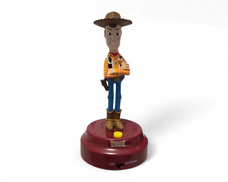 May include: A Woody action figure from the Toy Story franchise. The figure is wearing a cowboy hat, a yellow plaid shirt, blue pants, and brown boots. The figure is standing on a red base with the word "WOODY" and the words "ON" and "SENSOR".