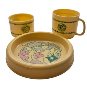 Könnte beinhalten: Ein Vintage-Babyservice in hellgelber Farbe. Das Set besteht aus einer runden Schüssel, einem Becher mit Henkel und einem kleinen Becher. Jedes Teil zeigt eine grün-rosa Illustration eines Babys und eines Storchs.