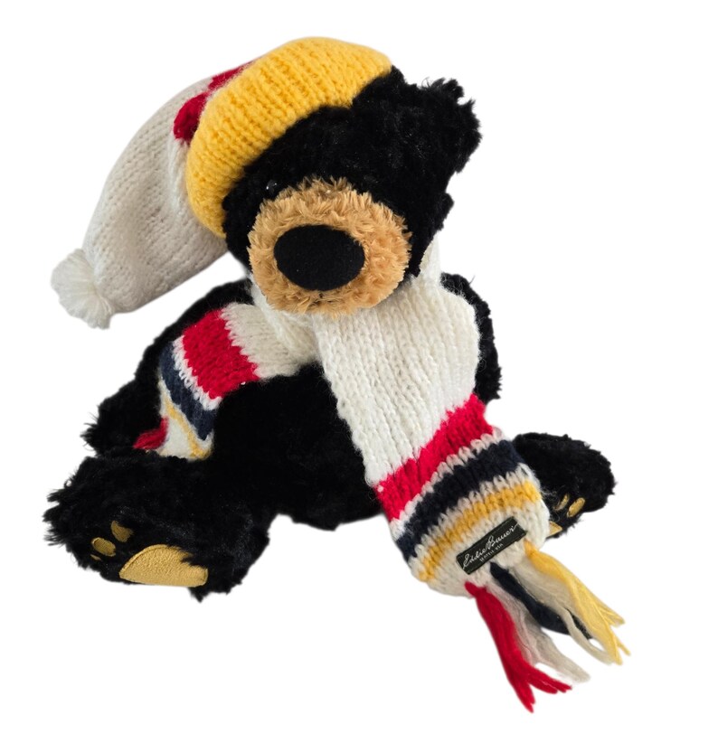 May include: A black plush bear wearing a white, red, and blue striped scarf and a knit cap with a yellow brim. The bear has a tan muzzle and yellow paw pads. The scarf has a tag that reads "Eddie Bauer".
