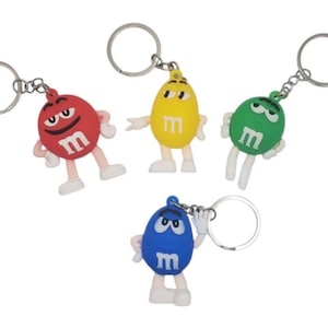 Puede incluir: Cuatro llaveros de personajes M&M's de colores. Los llaveros presentan personajes M&M's rojos, amarillos, verdes y azules con la letra "M" en sus cuerpos. Cada personaje tiene un llavero plateado.