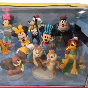 May include: A collection of Disney character figurines, including Mickey Mouse, Minnie Mouse, Goofy, Pluto, and Chip and Dale. The figures are in a clear plastic display case, with vibrant colors and detailed designs. The Pluto figure has a sign that says "PLUTO".