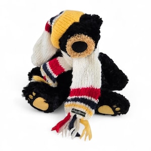 May include: A black plush teddy bear wearing a knit hat and scarf. The hat is yellow, white, and red striped. The scarf is white with red, navy, and yellow stripes. The bear has a brown nose and tan paws.