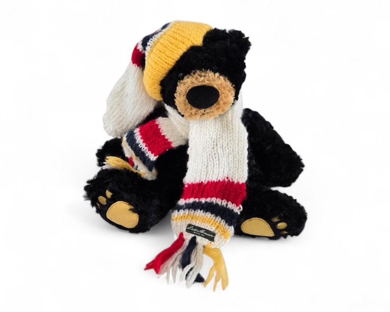 May include: A black plush teddy bear wearing a knit hat and scarf. The hat is yellow, white, and red striped. The scarf is white with red, navy, and yellow stripes. The bear has a brown nose and tan paws.