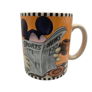 以下が含まれることがあります： 「SPORTS NEWS」の見出しが付いた新聞を読むネズミの漫画イラストが描かれたセラミックマグ。マグにはベージュのハンドルと白黒のチェック柄のリムが付いています。背景はオレンジ色です。