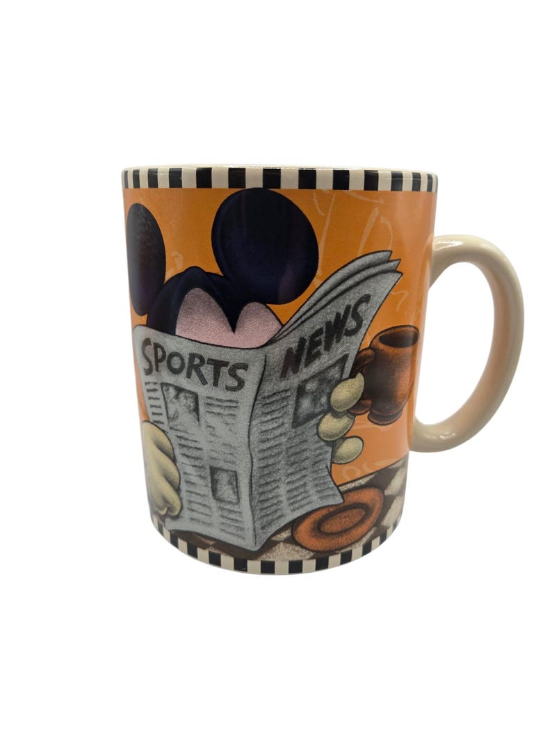 以下が含まれることがあります： 「SPORTS NEWS」の見出しが付いた新聞を読むネズミの漫画イラストが描かれたセラミックマグ。マグにはベージュのハンドルと白黒のチェック柄のリムが付いています。背景はオレンジ色です。