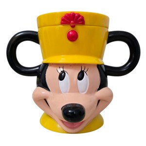 May include: A yellow and black Minnie Mouse-themed mug. The mug features Minnie's face, with black handles and a yellow hat with red accents. The mug is designed to resemble Minnie Mouse's iconic look.