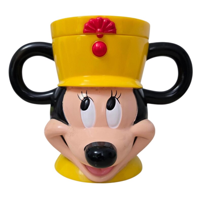 May include: A yellow and black Minnie Mouse-themed mug. The mug features Minnie's face, with black handles and a yellow hat with red accents. The mug is designed to resemble Minnie Mouse's iconic look.