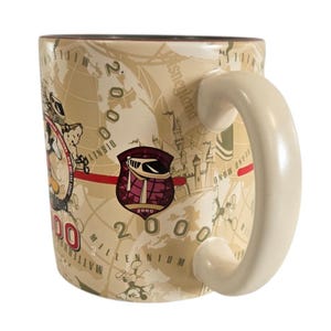 Pode incluir: Uma caneca de cer&acirc;mica bege com um design de mapa e o texto "Disneyland Millennium 2000". A caneca tem uma al&ccedil;a branca e um emblema bord&ocirc; e branco com o n&uacute;mero 2000.