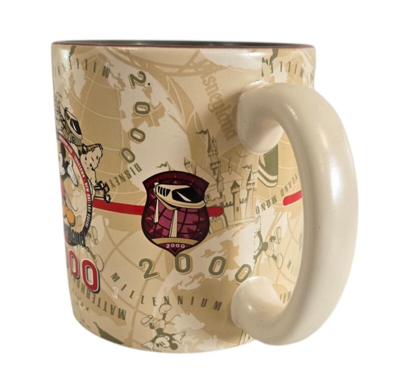 Pode incluir: Uma caneca de cer&acirc;mica bege com um design de mapa e o texto "Disneyland Millennium 2000". A caneca tem uma al&ccedil;a branca e um emblema bord&ocirc; e branco com o n&uacute;mero 2000.