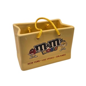 Puede incluir: Un recipiente de cerámica amarillo con forma de bolsa de la compra con un asa de cuerda amarilla. La parte delantera presenta el logotipo "M&M's World" con las ubicaciones Nueva York, Las Vegas y Orlando.