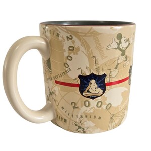 Pode incluir: Caneca de cer&acirc;mica bege com Mickey Mouse e um design de mapa-m&uacute;ndi. A caneca tem uma al&ccedil;a creme e um interior verde escuro. A caneca &eacute; decorada com o texto "2000 Millennium Disneyland Resort".