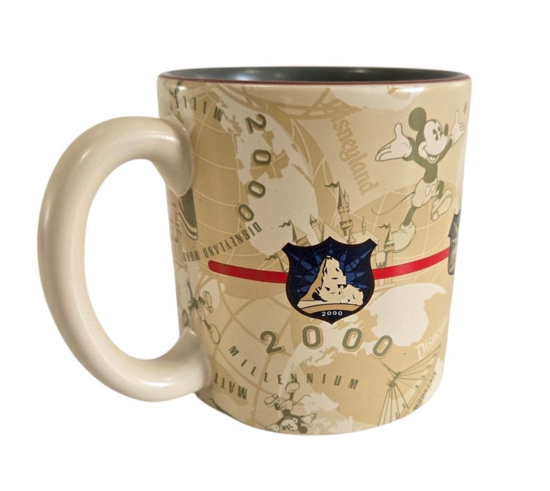 Pode incluir: Caneca de cer&acirc;mica bege com Mickey Mouse e um design de mapa-m&uacute;ndi. A caneca tem uma al&ccedil;a creme e um interior verde escuro. A caneca &eacute; decorada com o texto "2000 Millennium Disneyland Resort".