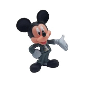 May include: A figurine of Mickey Mouse in a dark gray suit, white gloves, and black shoes. The iconic cartoon character has a pink face, black ears, and is posed with one arm outstretched.