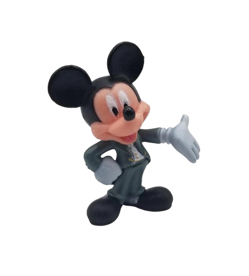 May include: A figurine of Mickey Mouse in a dark gray suit, white gloves, and black shoes. The iconic cartoon character has a pink face, black ears, and is posed with one arm outstretched.