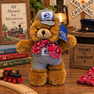 May include: A brown plush teddy bear wearing a blue and white striped engineer cap, matching striped overalls, and a red paisley bandana. The bear features a blue tag on its arm and a small patch on the chest, both displaying a train conductor graphic.