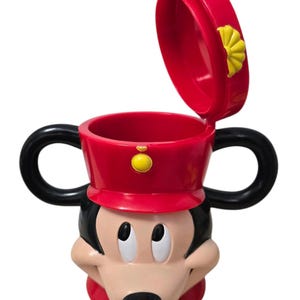 May include: A red and black Mickey Mouse-themed mug with a lid. The mug features Mickey's face, a red hat, and black handles. The lid is open, revealing the inside of the mug. A yellow button is on the hat.