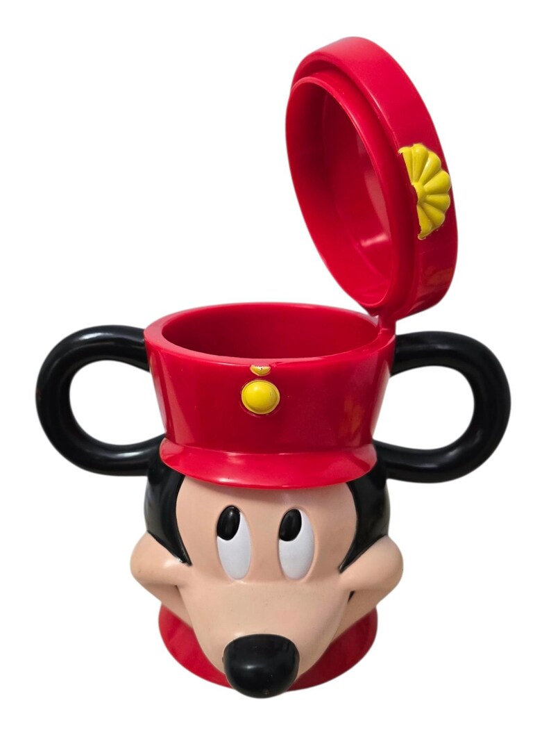 May include: A red and black Mickey Mouse-themed mug with a lid. The mug features Mickey's face, a red hat, and black handles. The lid is open, revealing the inside of the mug. A yellow button is on the hat.