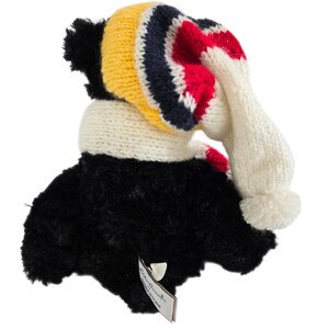 May include: A plush toucan toy with a black body and a knitted hat and scarf. The hat features yellow, navy, red, and white stripes. The scarf is white with red accents, and the hat has a white pom-pom.