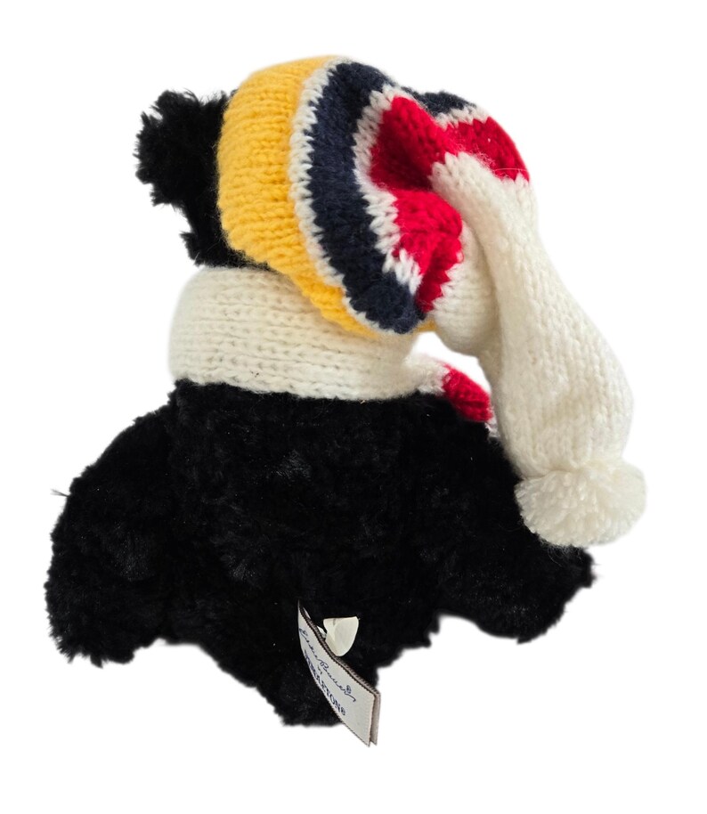May include: A plush toucan toy with a black body and a knitted hat and scarf. The hat features yellow, navy, red, and white stripes. The scarf is white with red accents, and the hat has a white pom-pom.