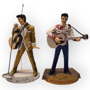 May include: Two figurines of a singer playing a guitar. One figure wears a gold suit and holds a microphone. The other figure wears a white shirt with a red pattern and blue trousers. Both figures are on a base with the singer's signature.