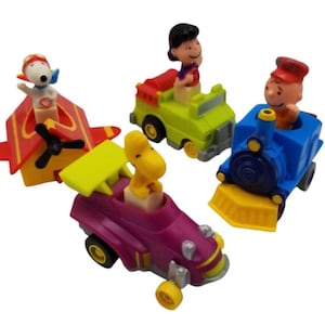 Op de afbeelding: Een verzameling kleurrijke speelgoedvoertuigen met personages uit de Peanuts-strip. Inclusief een rood en geel vliegtuig met Snoopy, een groene jeep met Lucy, een blauwe trein met Charlie Brown en een paarse raceauto met Woodstock.