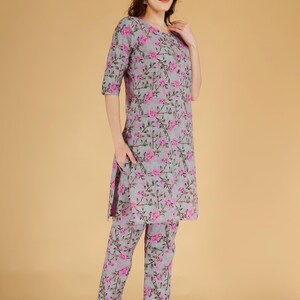 May include: A two-piece outfit comprising a grey tunic and matching trousers, both decorated with a pink floral design. The tunic has a round neckline and three-quarter sleeves. The trousers are straight-legged. The outfit is made of a lightweight fabric.
