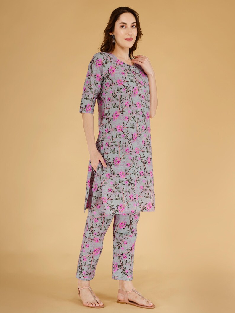 May include: A two-piece outfit comprising a grey tunic and matching trousers, both decorated with a pink floral design. The tunic has a round neckline and three-quarter sleeves. The trousers are straight-legged. The outfit is made of a lightweight fabric.