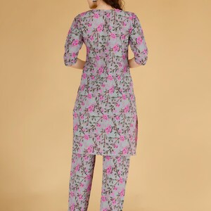 May include: A two-piece floral print outfit in grey and pink. The set includes a long-sleeved tunic and matching trousers. The floral pattern features pink flowers and green leaves on a grey background. The outfit is made of a lightweight fabric.