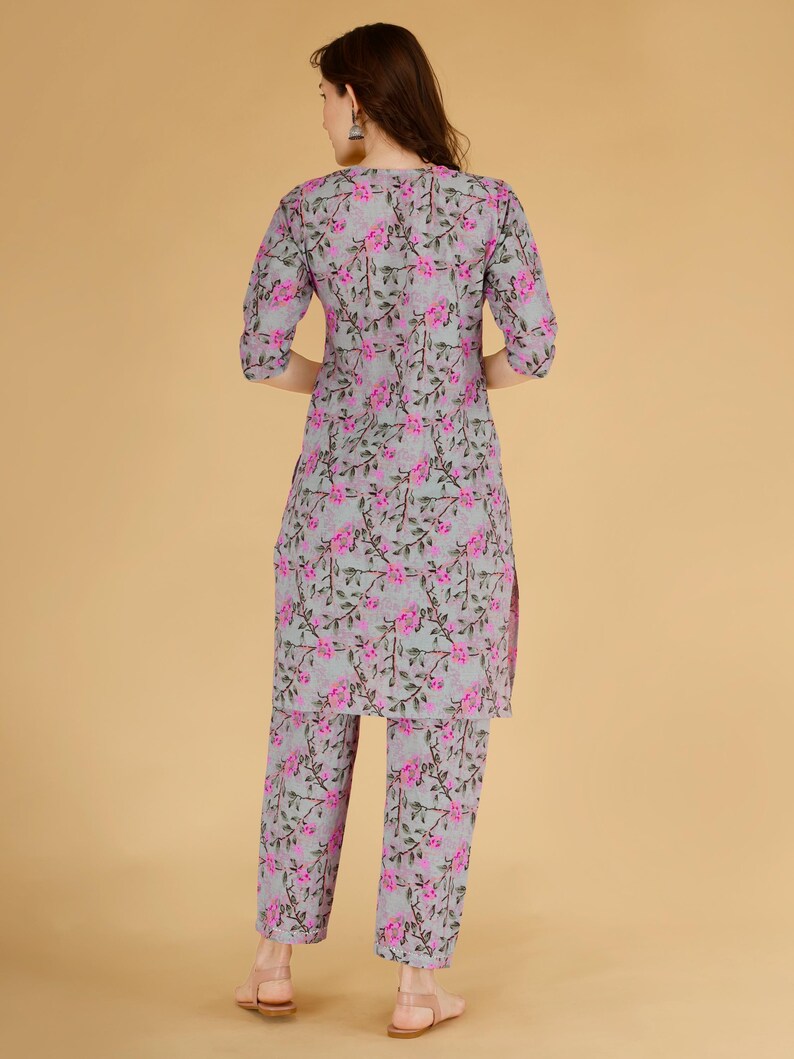May include: A two-piece floral print outfit in grey and pink. The set includes a long-sleeved tunic and matching trousers. The floral pattern features pink flowers and green leaves on a grey background. The outfit is made of a lightweight fabric.