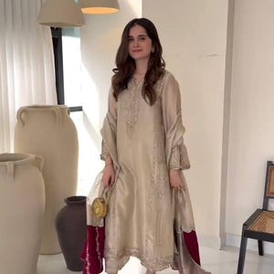 Puede incluir: Una mujer con un conjunto de seda beige con bordados detallados y mangas acampanadas. El conjunto incluye pantalones a juego y un chal transparente. Lleva un clutch dorado y sandalias de tacón alto. Adecuado para ocasiones especiales.