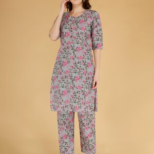 May include: A grey and pink floral two-piece outfit. The set comprises a tunic top with three-quarter sleeves and matching cropped trousers. The floral design features pink flowers and green stems.
