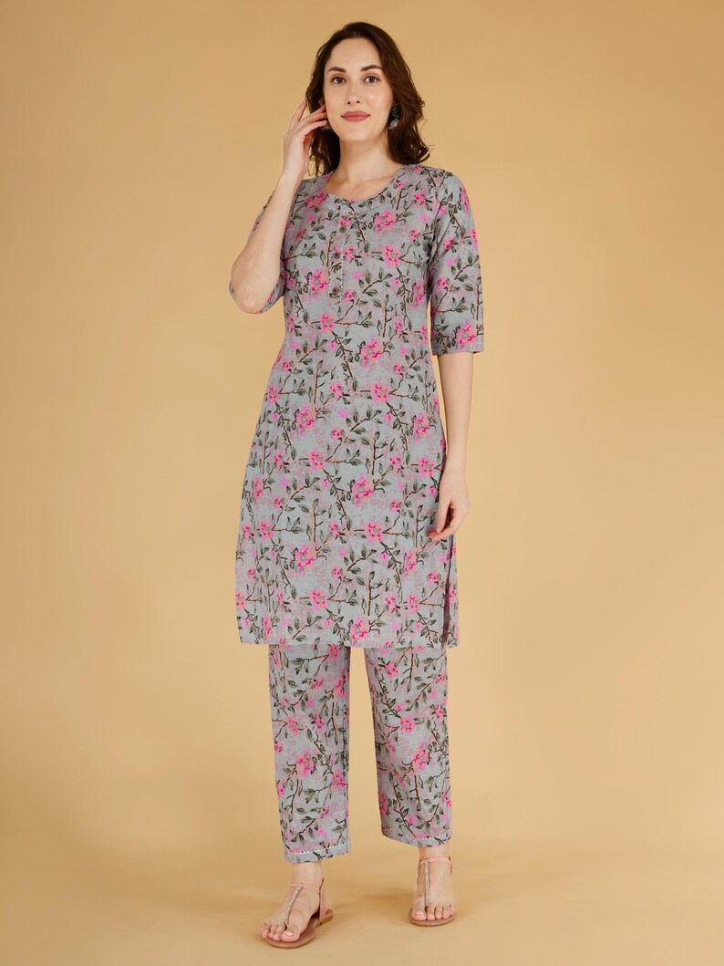 May include: A grey and pink floral two-piece outfit. The set comprises a tunic top with three-quarter sleeves and matching cropped trousers. The floral design features pink flowers and green stems.