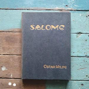 Könnte beinhalten: Ein schwarzes Hardcover-Buch mit dem Titel Salome von Oscar Wilde. Auf dem Vorderdeckel steht der Titel Salome in großen, stilisierten goldenen Buchstaben oben und der Autorenname Oscar Wilde in kleineren goldenen Buchstaben unten rechts.