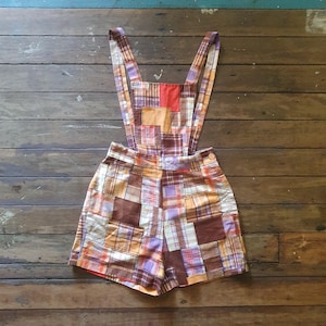 May include: Patchwork overall shorts with a brown, orange and purple plaid pattern. The straps are wide and have a simple design.