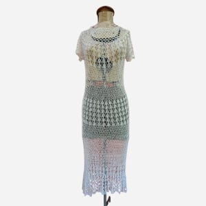 K&ouml;nnte beinhalten: Wei&szlig;es H&auml;kelkleid mit offenem Gewebe, kurzen &Auml;rmeln und Midi-L&auml;nge. Das Kleid wird auf einer Schaufensterpuppe pr&auml;sentiert, die das komplizierte Muster und die zarte Textur zeigt. Das Kleid hat einen gewellten Saum.